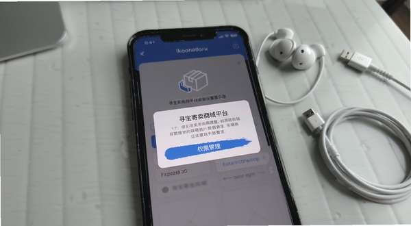 寻宝寄卖商城平台安装设置界面,画面主体为手机屏幕中的权限管理弹窗,背景区域为浅灰色桌面,旁边放置耳机与数据线,整体风格生活化
